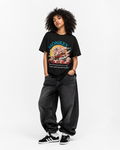 A model wearing a black graphic t-shirt with a colorful monkey print, paired with dark gray baggy jeans and black and white sneakers, standing against a plain white studio background.