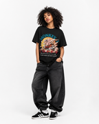 A model wearing a black graphic t-shirt with a colorful monkey print, paired with dark gray baggy jeans and black and white sneakers, standing against a plain white studio background.