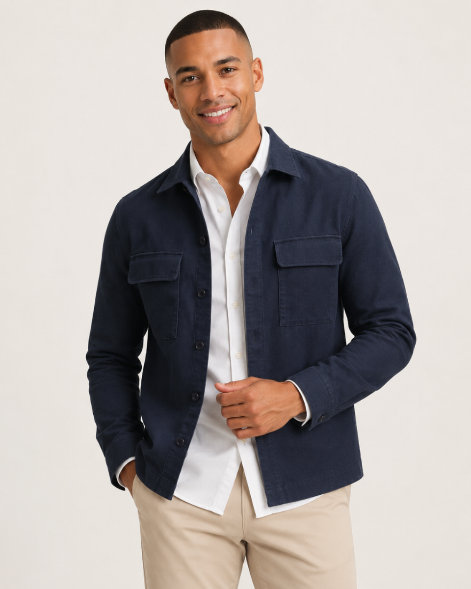 A man wearing a navy blue button-up overshirt over a white collared shirt and beige trousers, standing against a light gray studio background.