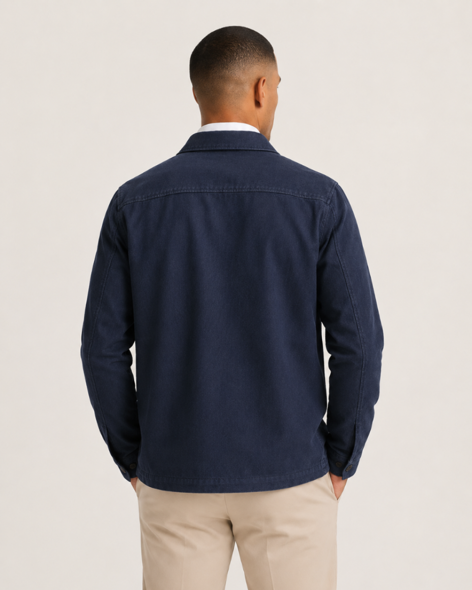 A man viewed from the back wearing a navy blue overshirt or lightweight jacket over a white collared shirt and beige trousers, against a plain light gray studio background.