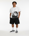 A man modeling a white graphic t-shirt with a black anime-style print, paired with black shorts, white socks, and black-and-white sneakers, standing against a textured gray wall.