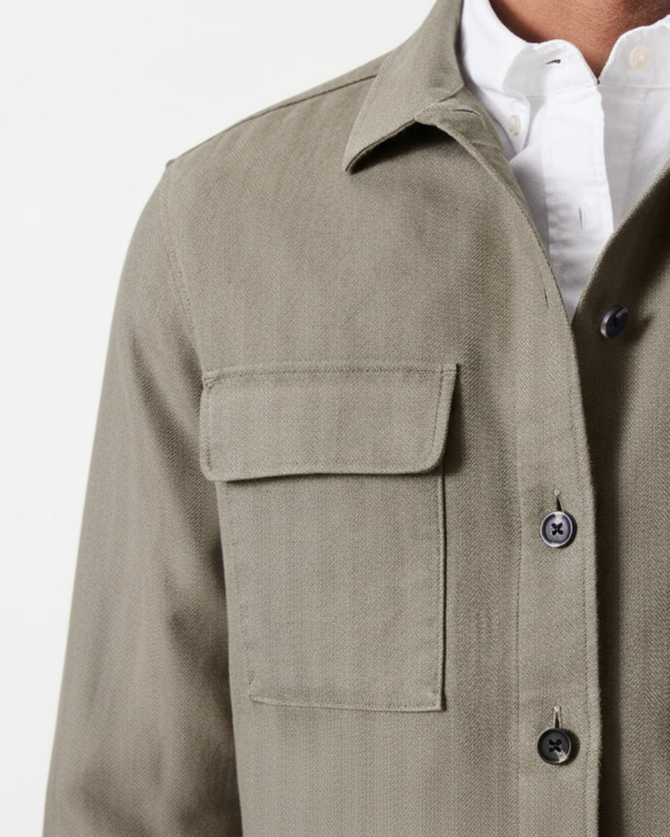 Close-up of a person wearing a textured light brown linen button-up shirt jacket over a white collared shirt, showing the chest pocket and fabric texture.