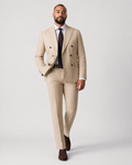 A man modeling a light beige double-breasted suit with a white dress shirt, dark tie, and brown shoes against a light gray studio background.