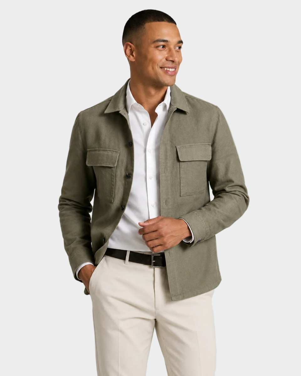 A man wearing a slim-fit olive green button-up shirt jacket over a white collared shirt, paired with off-white trousers and a black belt, photographed against a light gray studio background.