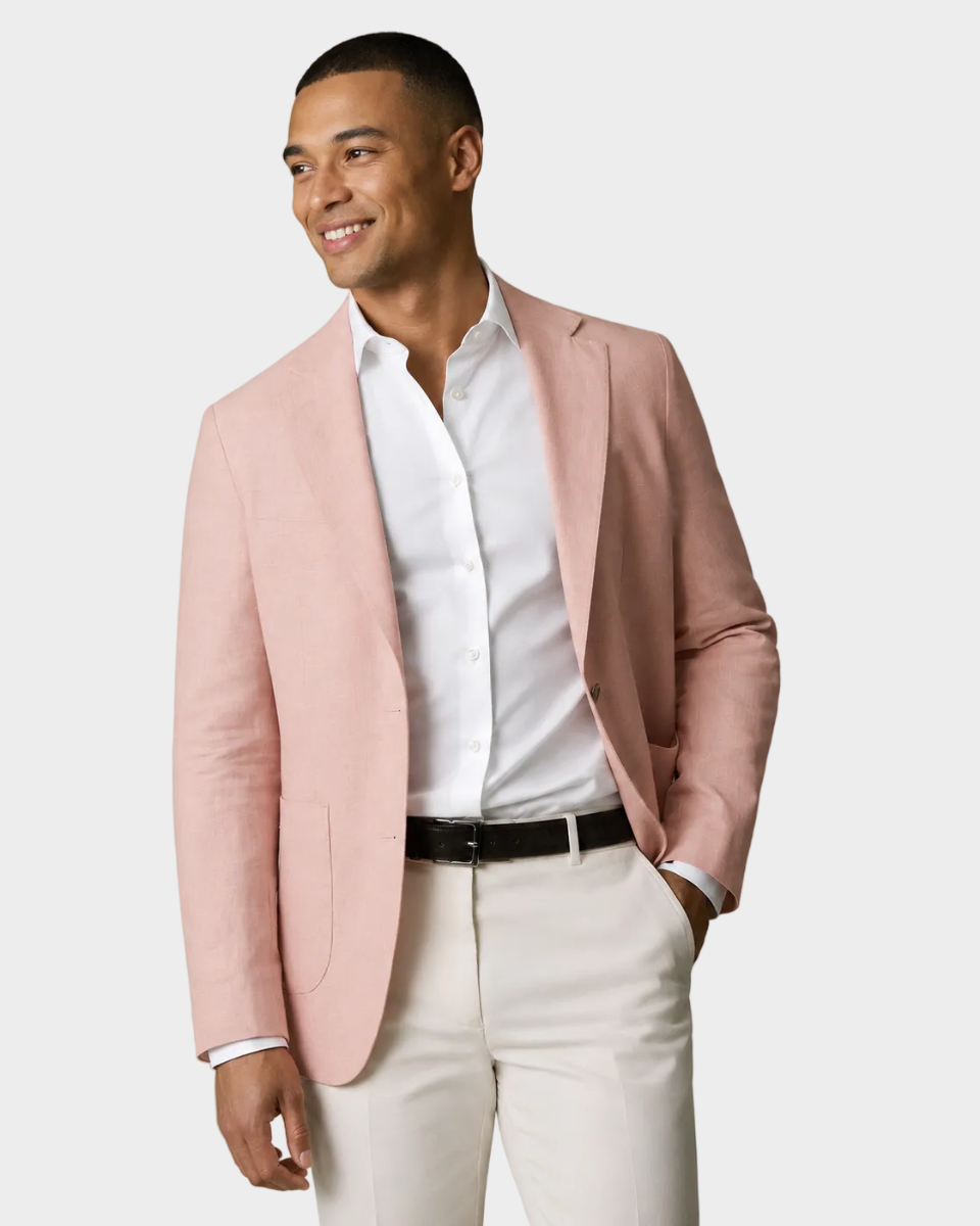A man wearing a light pink blazer over a white dress shirt and cream trousers, standing against a plain beige studio background.