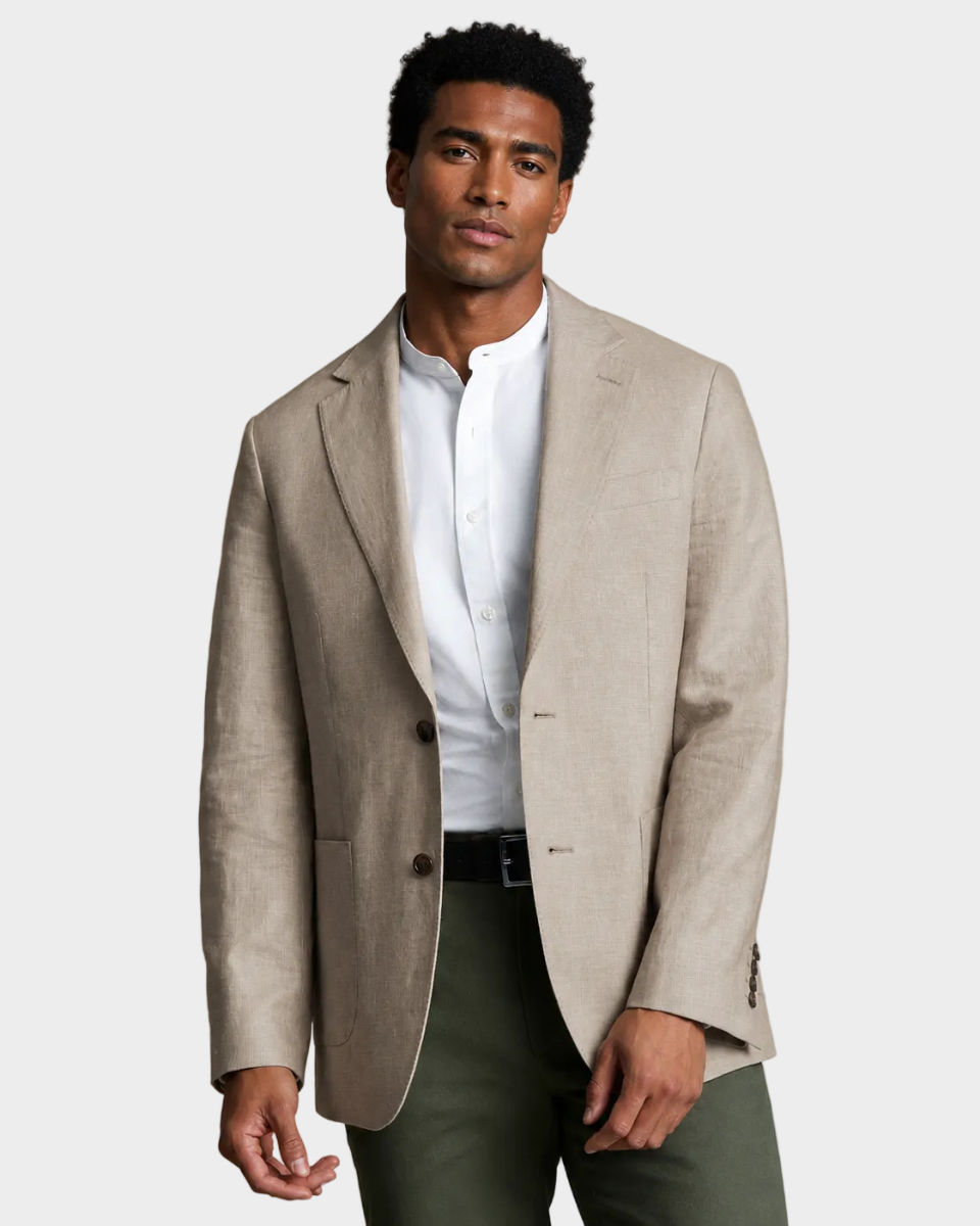 A man wearing a light beige linen-blend blazer over a white mandarin-collar shirt, paired with dark olive trousers, photographed against a solid warm beige studio background.