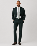 A man modeling a dark forest green two-piece suit with a white dress shirt, a light gray tie, and dark brown leather dress shoes, standing against a light gray studio background.