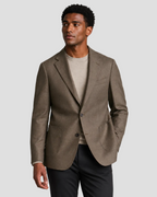 A man wearing a brown textured blazer over a beige crewneck sweater and dark trousers, standing against a light gray studio background.