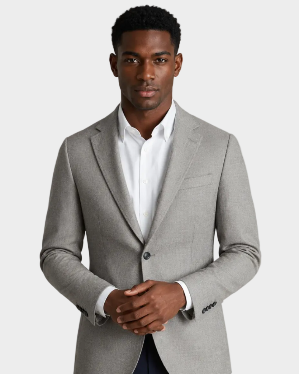 A man wearing a light gray tailored blazer over a white dress shirt and navy trousers, standing against a plain beige studio background.
