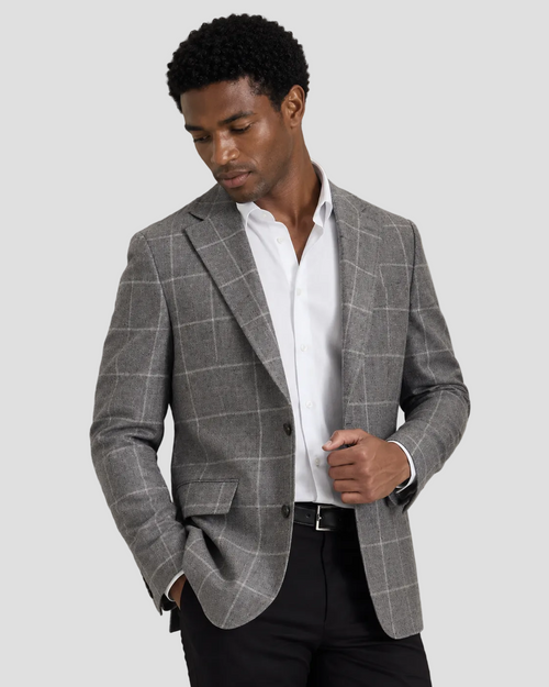 A man wearing a gray windowpane plaid blazer over a white dress shirt and black trousers, standing against a light gray studio background.