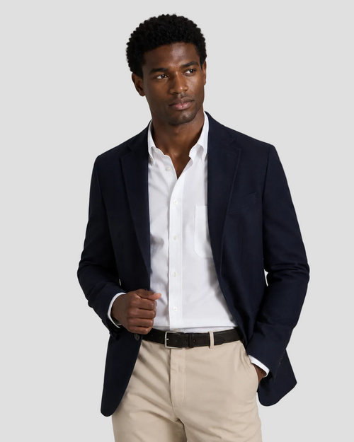 A man wearing a navy blue blazer over a white dress shirt, paired with beige trousers and a black belt, standing against a neutral beige studio background.