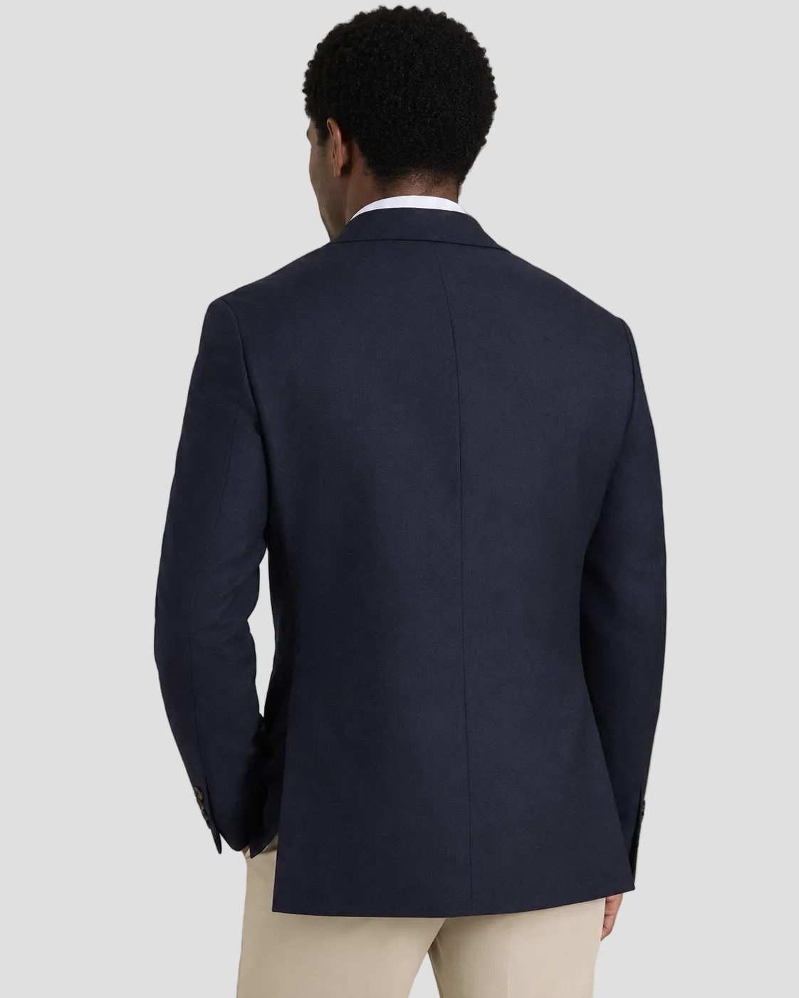 A man viewed from the back wearing a dark navy blue blazer over a white collared shirt and light beige trousers, set against a plain beige studio background.