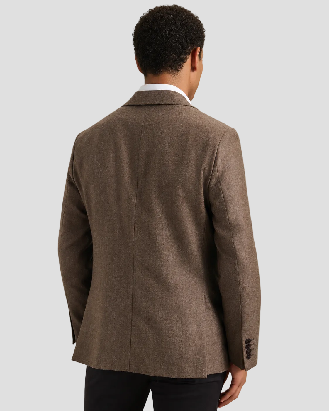 A man viewed from the back wearing a brown textured blazer over a white collared shirt and dark trousers, set against a plain light gray studio background.