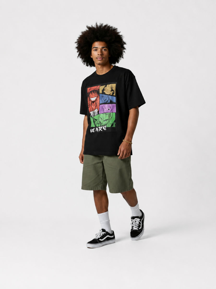 A model with an afro hairstyle stands against a light gray studio background wearing a black graphic t-shirt, olive green shorts, white socks, and black Vans sneakers. The t-shirt features a colorful comic-style graphic of Marvel characters with the text 'WE ARE'.