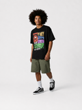 A model with an afro hairstyle stands against a light gray studio background wearing a black graphic t-shirt, olive green shorts, white socks, and black Vans sneakers. The t-shirt features a colorful comic-style graphic of Marvel characters with the text 'WE ARE'.