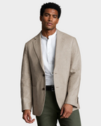 A man wearing a light beige linen-blend blazer over a white mandarin-collar shirt, paired with dark olive trousers, photographed against a solid warm beige studio background.
