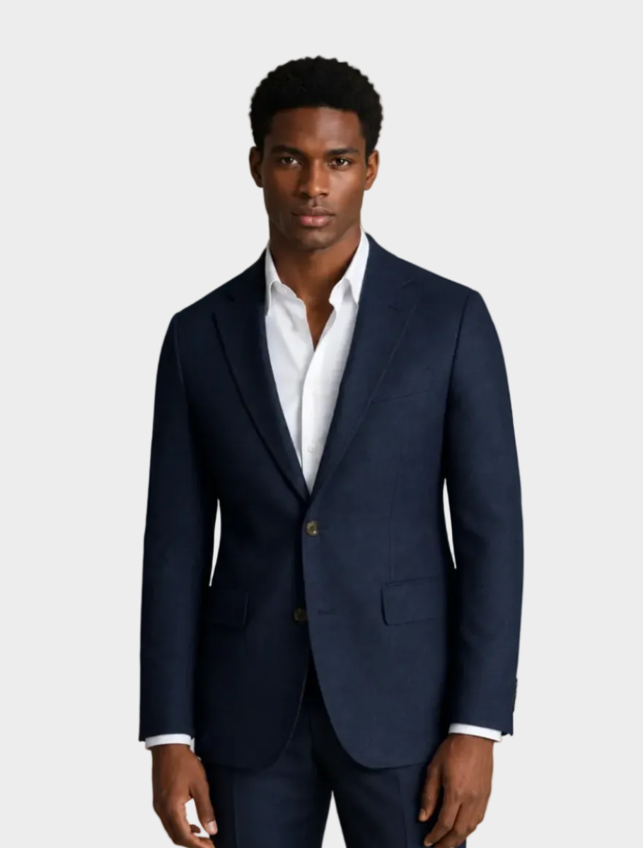 A man wearing a navy blue two-button suit jacket with a white dress shirt, standing against a solid beige background.