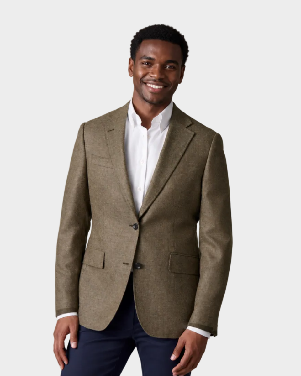 A man wearing a brown textured blazer over a white dress shirt and navy trousers, standing against a plain beige studio background.