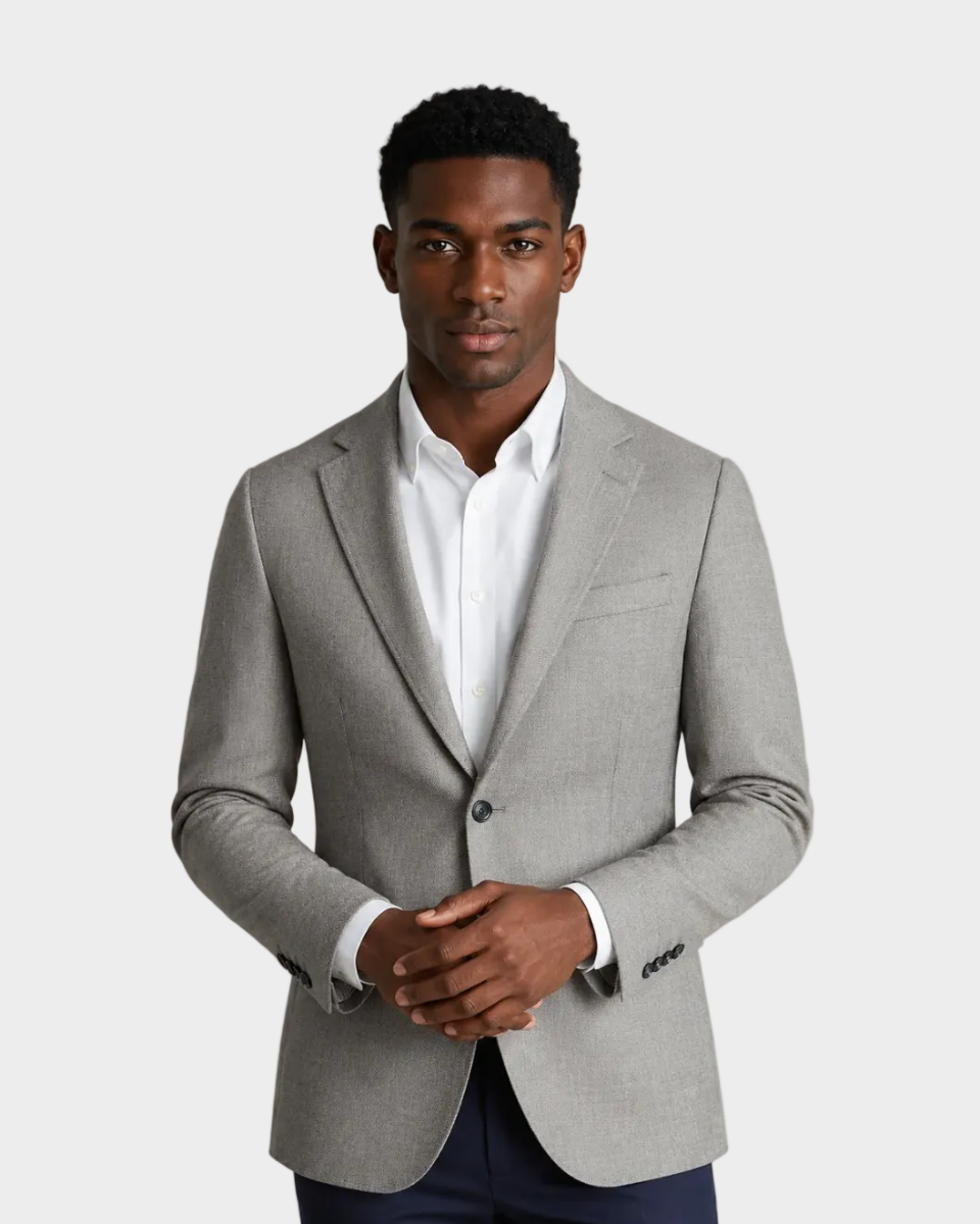 A man wearing a light gray tailored blazer over a white dress shirt and navy trousers, standing against a plain beige studio background.