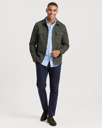 A man modeling a dark olive green utility jacket over a light blue button-down shirt, paired with navy trousers and brown suede shoes, against a plain light gray studio background.
