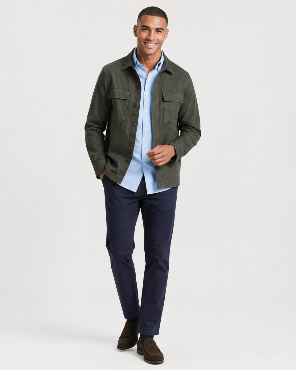 A man modeling a dark olive green utility jacket over a light blue button-down shirt, paired with navy trousers and brown suede shoes, against a plain light gray studio background.