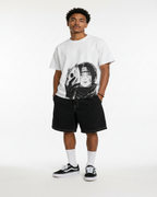 A man modeling a white graphic t-shirt with a black anime-style print, paired with black shorts, white socks, and black-and-white sneakers, standing against a textured gray wall.
