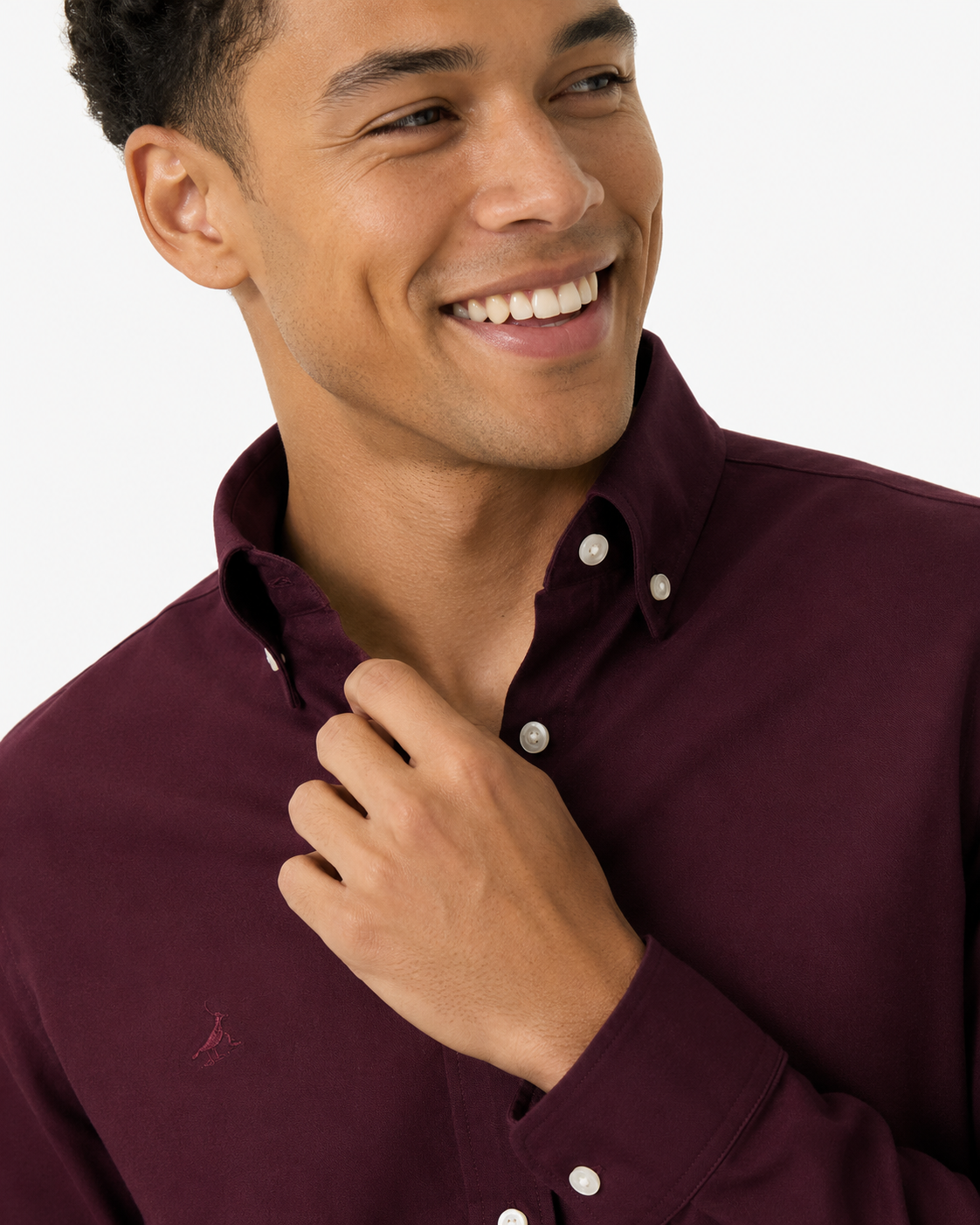 A close-up of a smiling man wearing a burgundy button-down shirt with white buttons and a small embroidered logo on the chest, set against a plain light background.