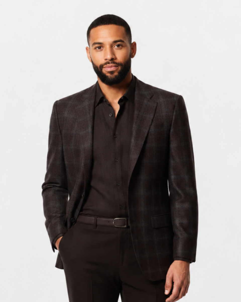 A man wearing a dark brown plaid blazer over a black shirt and black trousers, standing against a light gray studio background.