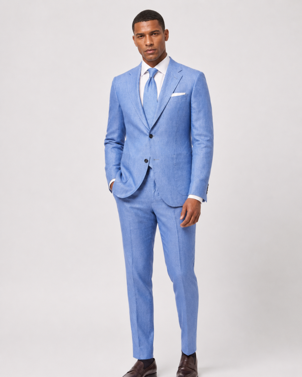 A man modeling a light blue textured two-piece suit with a white dress shirt, light blue tie, and white pocket square, paired with dark brown leather dress shoes, against a light gray studio background.