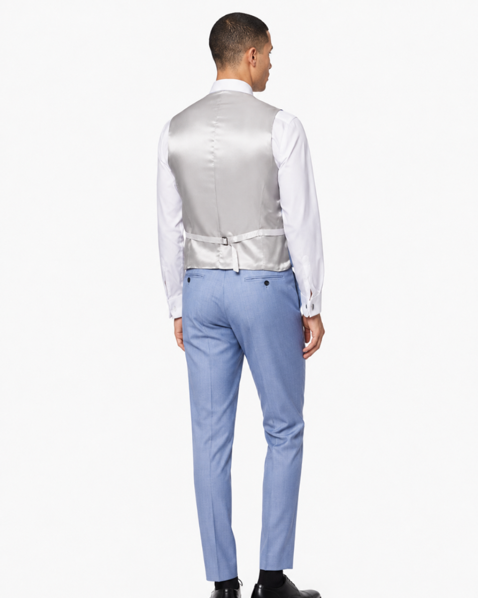 A man viewed from the back wearing a white dress shirt, a light silver satin vest, and light blue trousers, standing against a plain white studio background.
