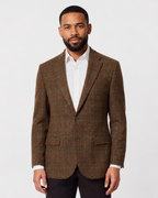 A man wearing a brown plaid blazer over a white collared shirt and dark trousers, standing against a light gray studio background.