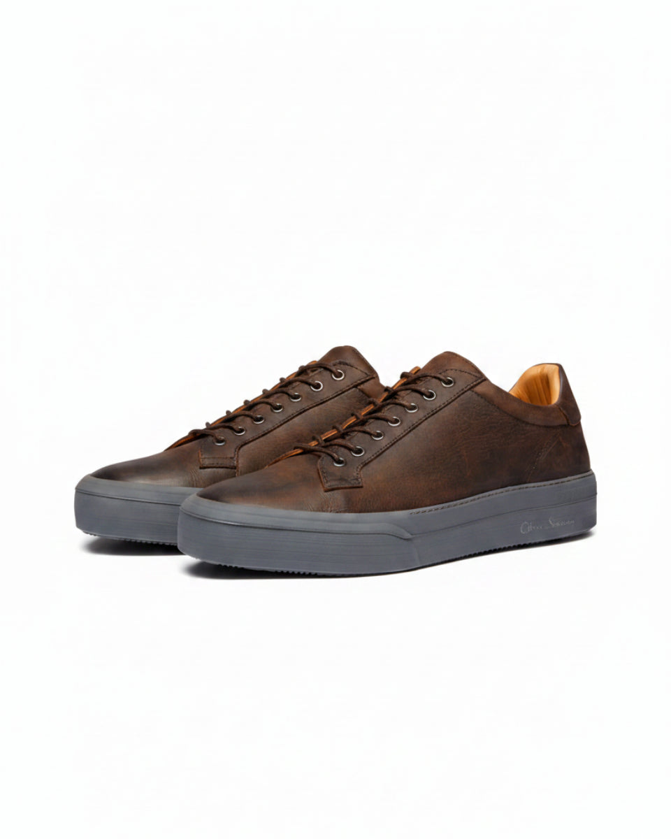 A pair of low-top leather sneakers in dark brown with matching brown laces, featuring a thick dark gray sole and a light tan interior lining. The brand name 'Calvin Klein' is subtly printed on the side of the gray sole.