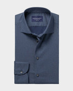 A folded navy blue dress shirt with a small white geometric pattern, featuring a pointed collar, button-down front, and a single cuff with a button closure. The shirt is presented on a solid light beige background.