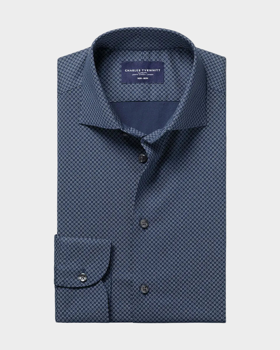A folded navy blue dress shirt with a small white geometric pattern, featuring a pointed collar, button-down front, and a single cuff with a button closure. The shirt is presented on a solid light beige background.