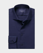 A folded navy blue dress shirt with a subtle micro-pattern, featuring a pointed collar, button-down front, and a single-button cuff, presented on a solid light beige background.