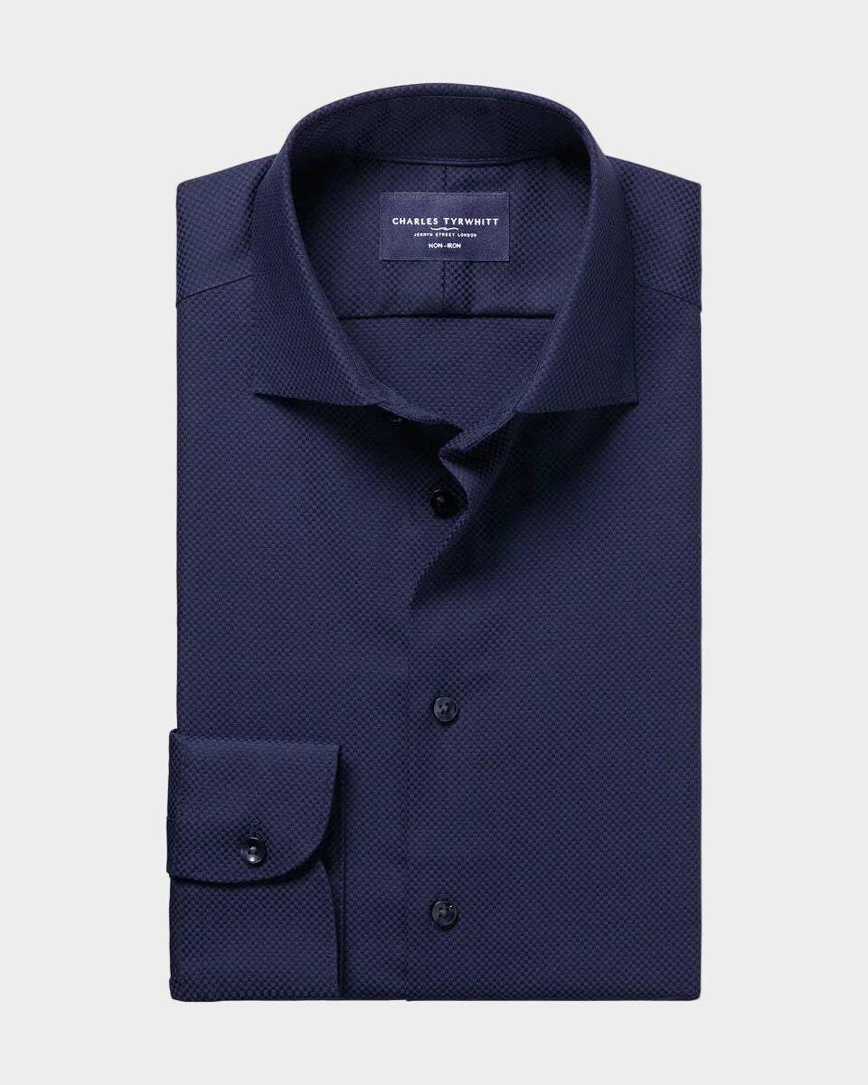 A folded navy blue dress shirt with a subtle micro-pattern, featuring a pointed collar, button-down front, and a single-button cuff, presented on a solid light beige background.