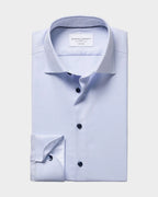 A light blue dress shirt folded neatly, showing a pointed collar, button front, and patterned inner placket and cuff, photographed on a plain warm beige background. The neck label reads Charles Tyrwhitt