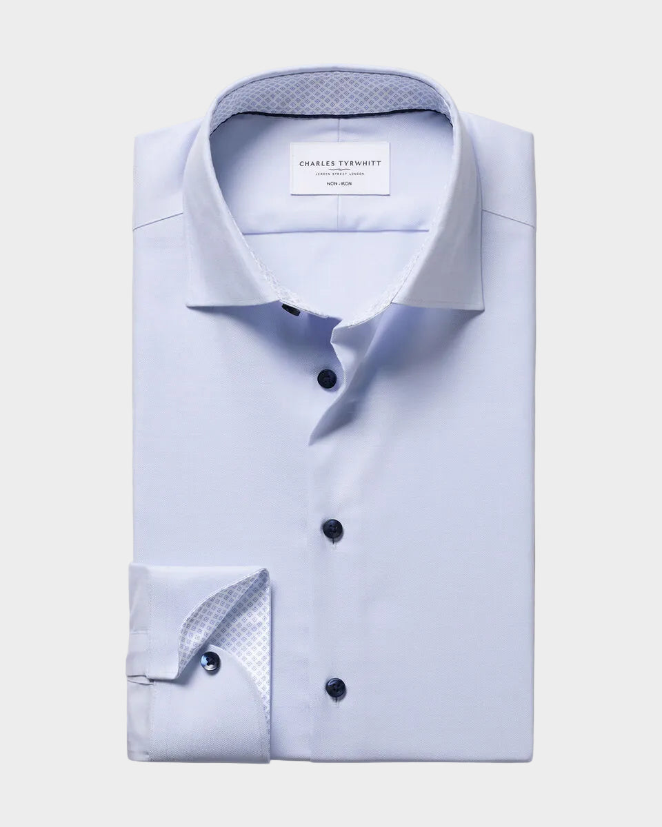 A light blue dress shirt folded neatly, showing a pointed collar, button front, and patterned inner placket and cuff, photographed on a plain warm beige background. The neck label reads Charles Tyrwhitt