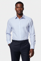 A man wearing a light blue long-sleeve button-down dress shirt and dark trousers, standing against a light gray studio background.