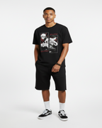 A man modeling a black graphic t-shirt and matching black shorts, styled with white socks and black-and-white sneakers, standing against a textured gray wall.