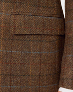 Close-up of a brown tweed blazer fabric with a subtle windowpane plaid pattern and a flap pocket, showing a hint of a white shirt cuff.