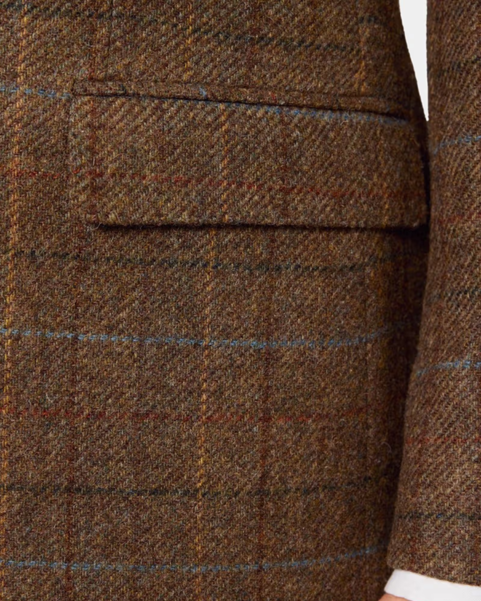 Close-up of a brown tweed blazer fabric with a subtle windowpane plaid pattern and a flap pocket, showing a hint of a white shirt cuff.