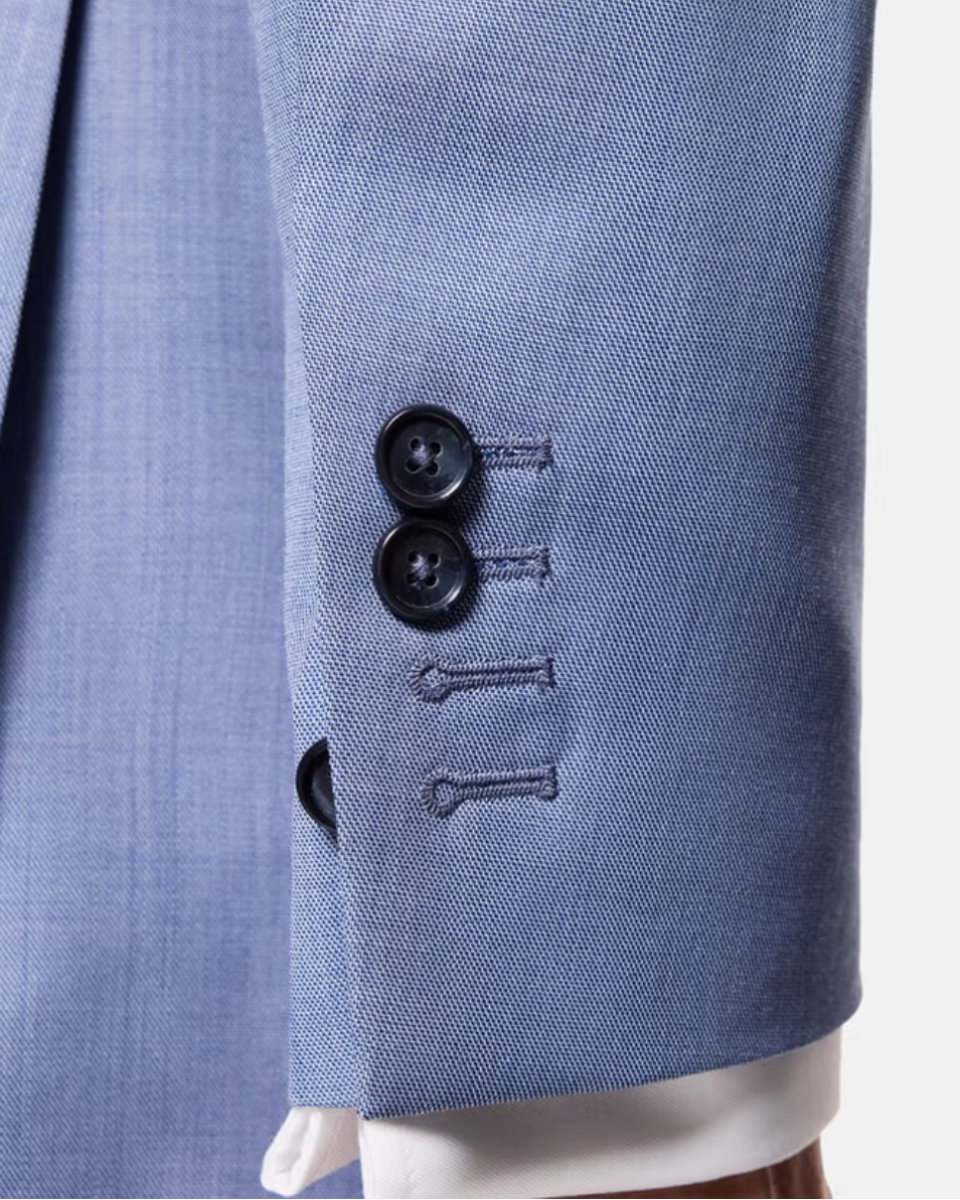 Close-up of a light blue textured suit jacket sleeve with two black buttons and decorative embroidered details, shown with a white dress shirt cuff.