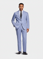 A man modeling a light blue three-piece suit with a white dress shirt, black tie, and black dress shoes against a light gray studio background.