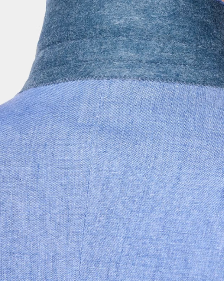 Close-up back view of a person wearing a light blue textured shirt with a contrasting darker blue high collar, against a plain light background.