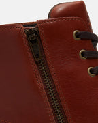 Close-up of a brown leather lace-up boot showing the side zipper and metal eyelets with dark brown laces against a light gray background.