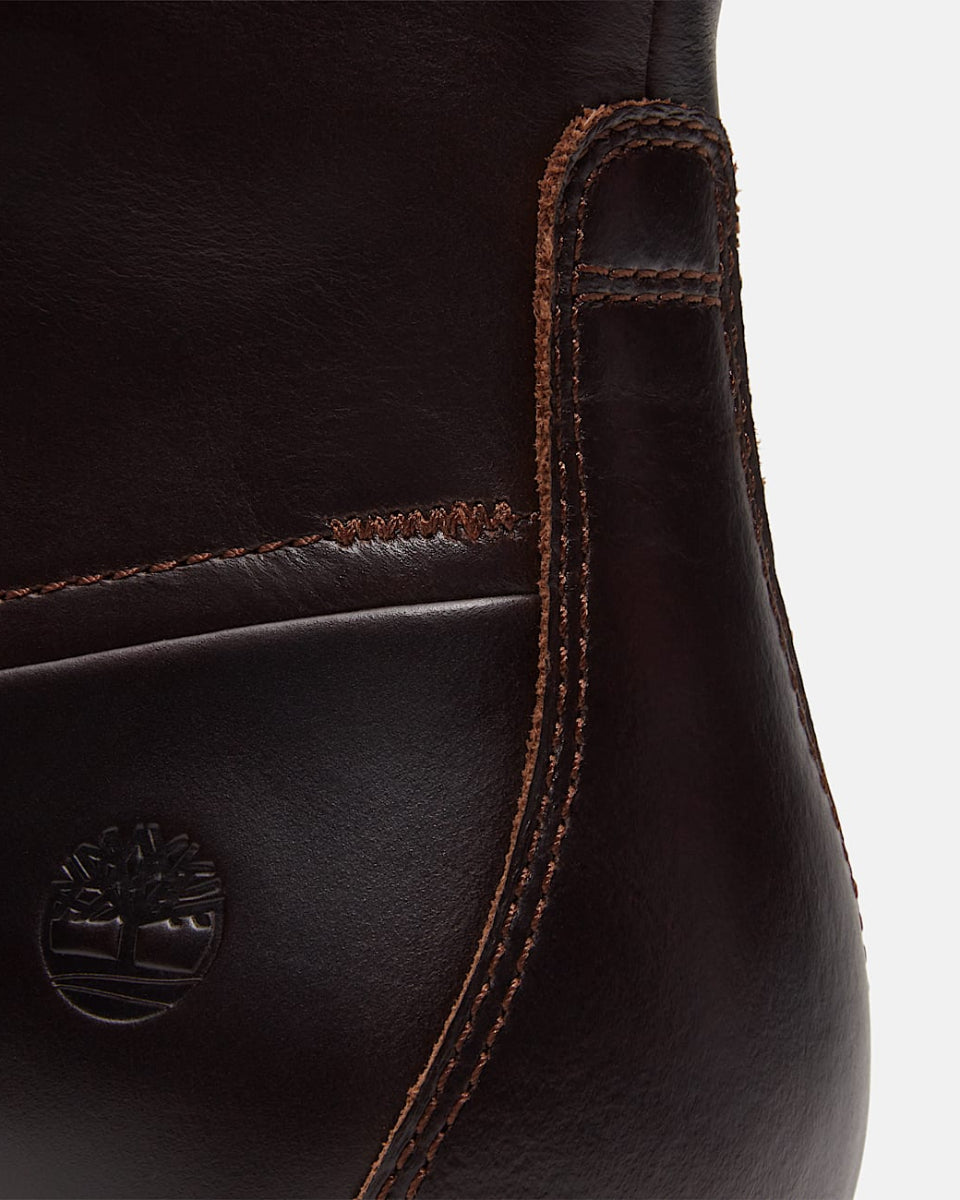 Close-up of a dark brown leather boot showing the side seam, contrast stitching, and an embossed Timberland tree logo on a light gray background.