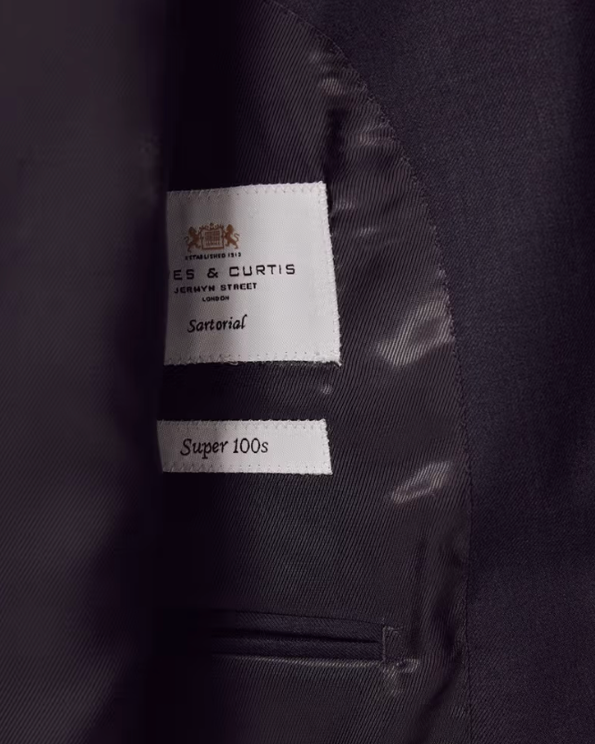 Close-up of the interior lining of a dark suit jacket, showing a white fabric label that reads ES & CURTIS and Sartorial, and another label reading Super 100s.